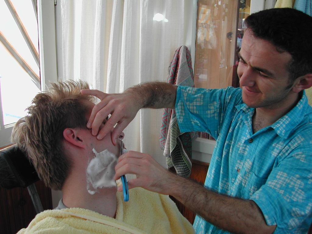 A barber in a blue shirt shaves a customer's face with a razor while holding their skin taut.