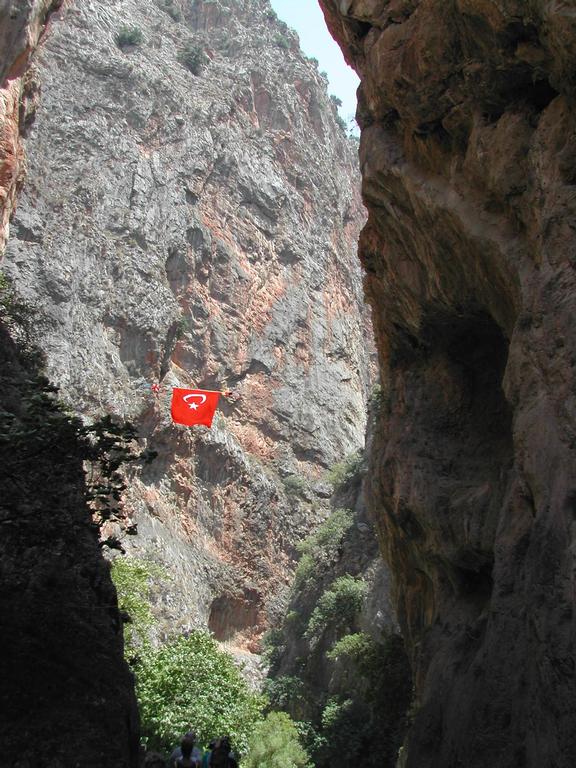 A red Turkish flag hangs between steep canyon walls, with people walking below among green vegetation.