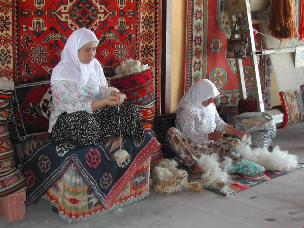 Two women in headscarves spin and prepare wool for weaving, sitting among colorful carpets and textiles.