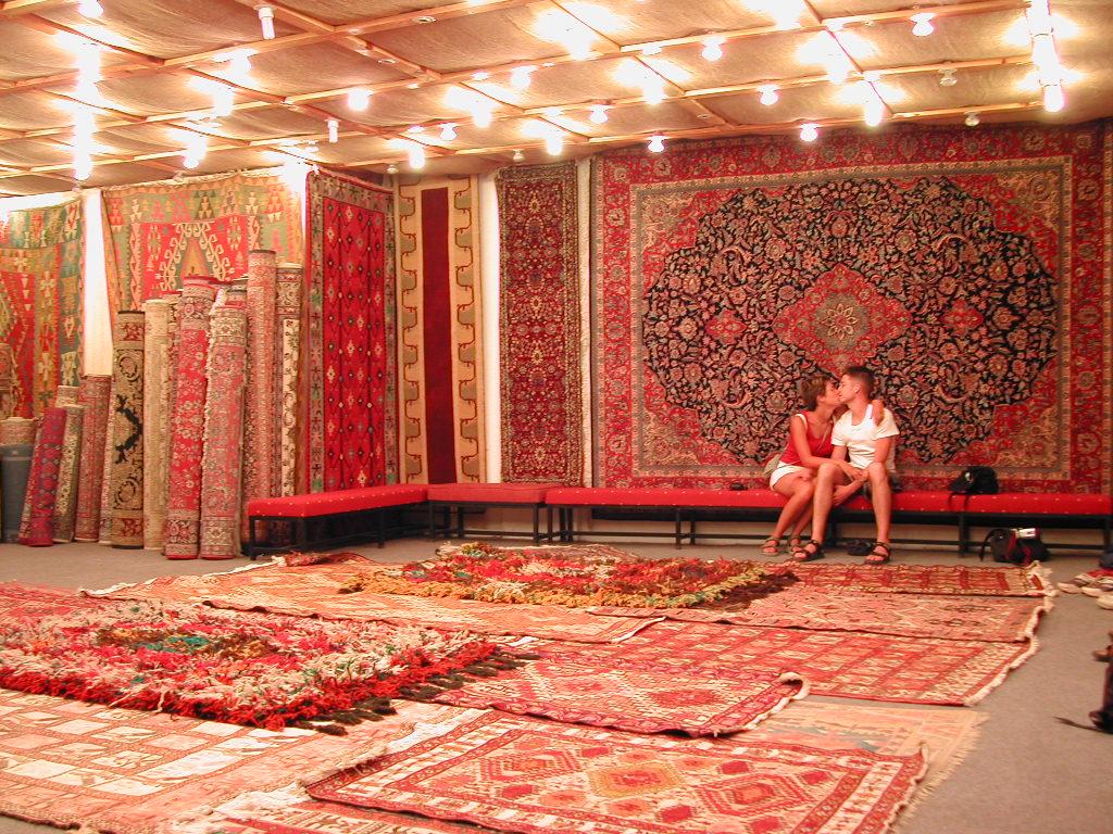 A room filled with colorful carpets and rugs, with a couple sitting on a bench embracing.