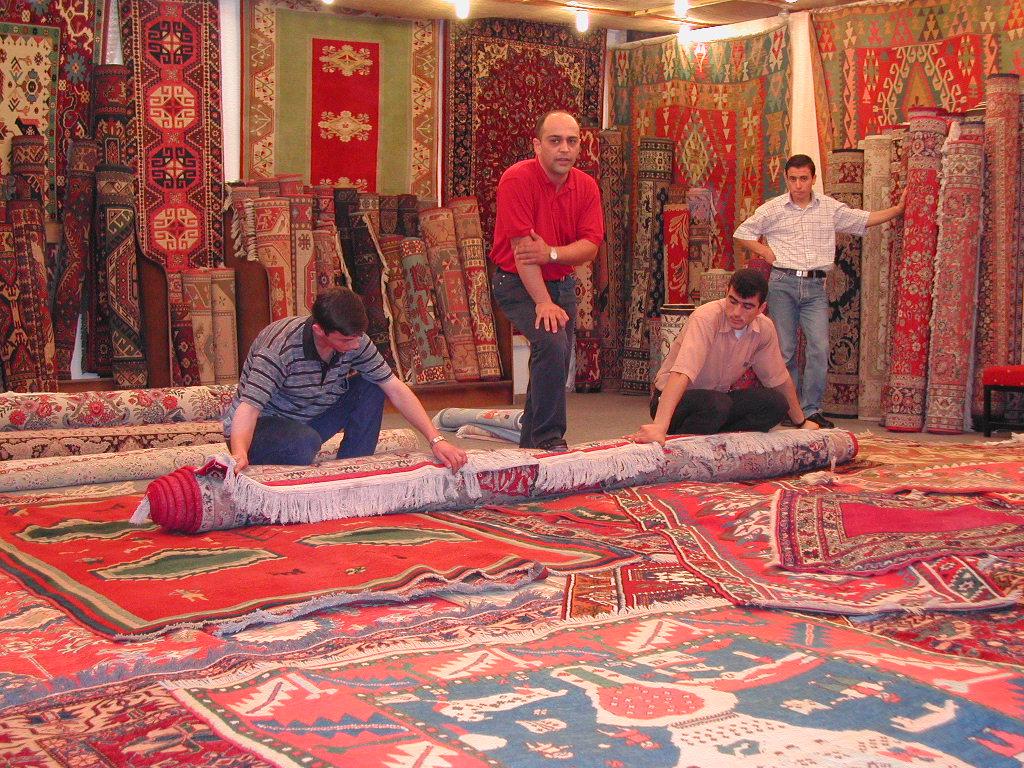 Three men unroll a large, patterned carpet in a shop, while another man stands among stacked rugs in the background.