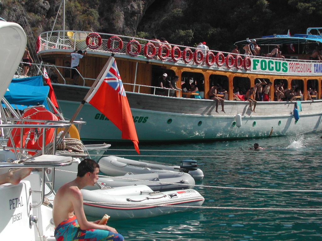 A large tour boat with people on board and swimming nearby, while a man eats on a smaller boat.