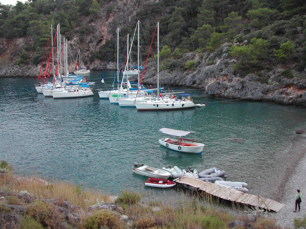 Several sailboats are anchored in a calm bay near a rocky shoreline, with small boats docked at a wooden pier.