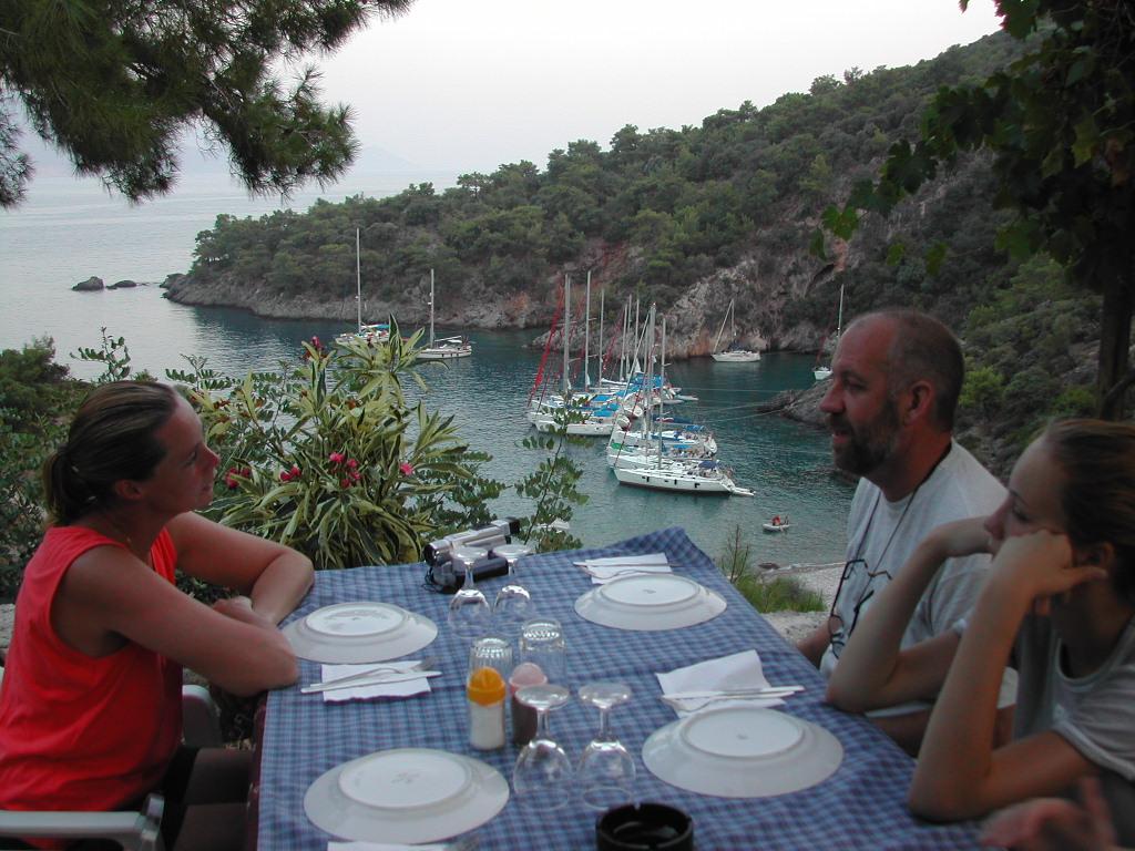 Three people sit at a table with a blue checkered tablecloth, overlooking a bay with anchored sailboats.