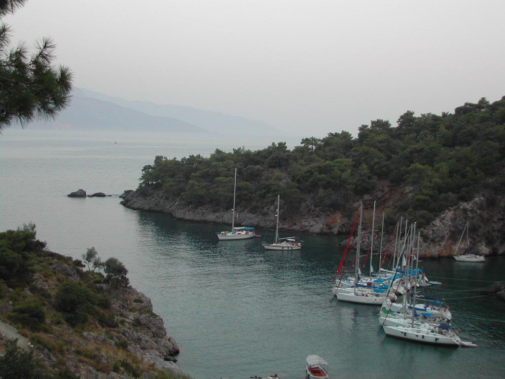 Several sailboats are anchored in a calm bay surrounded by rocky cliffs and green trees.