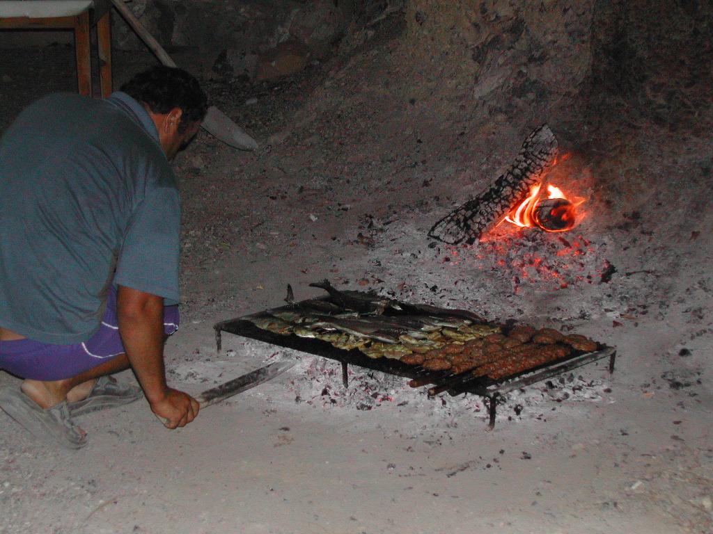 A man crouches near an open fire, grilling fish and meat on a metal grate over hot coals.