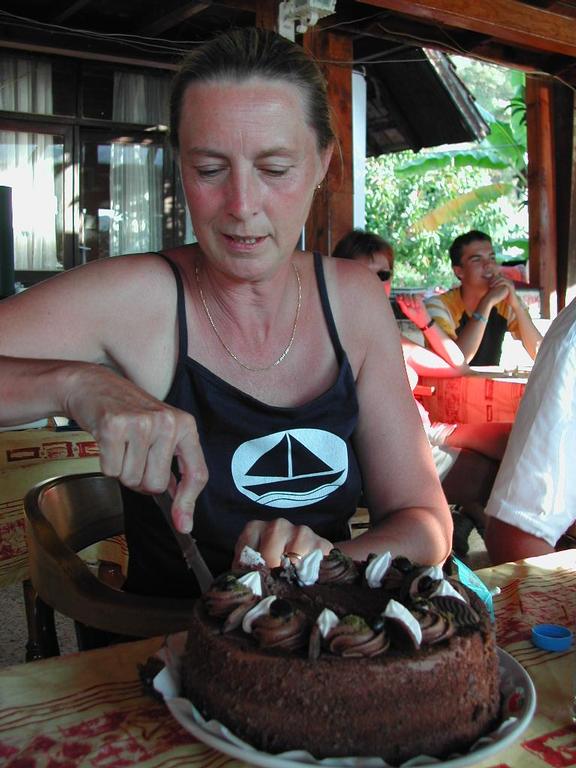 A woman in a black tank top cuts a chocolate cake at an outdoor table with people in the background.