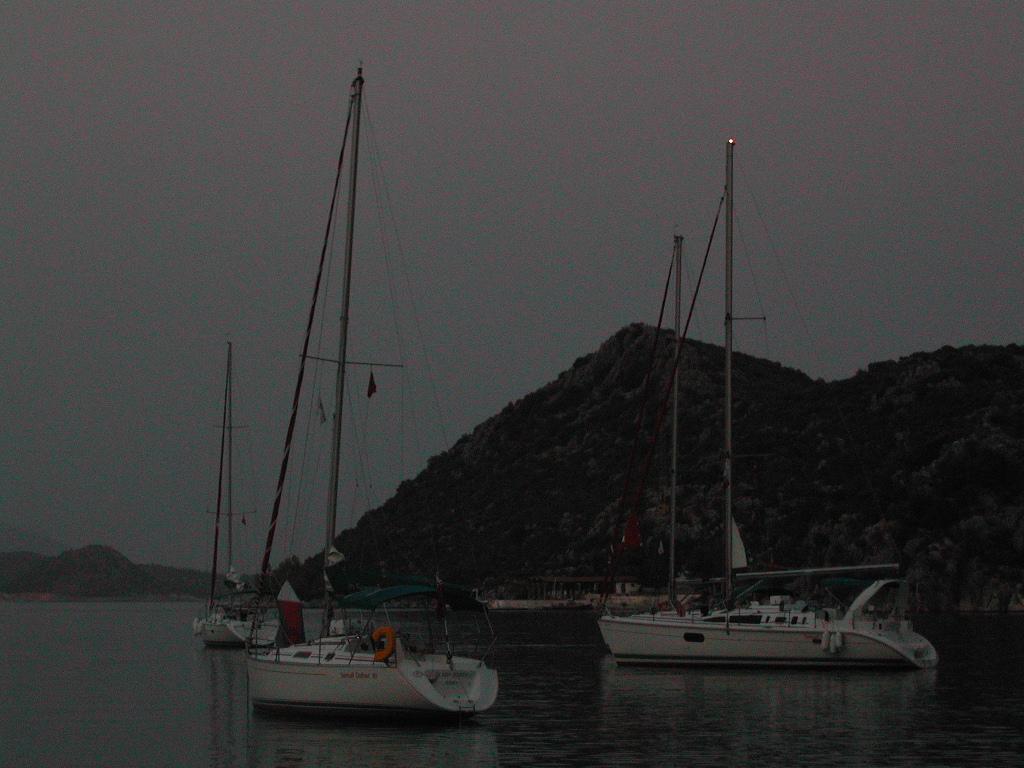 Several sailboats anchored in calm water near a rocky hillside, with navigation lights visible in the dim lighting.