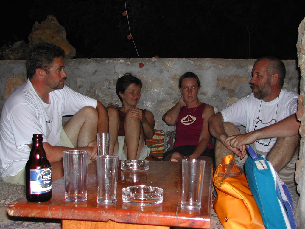 Four people sit around a wooden table with drinks, engaged in conversation in an outdoor setting at night.