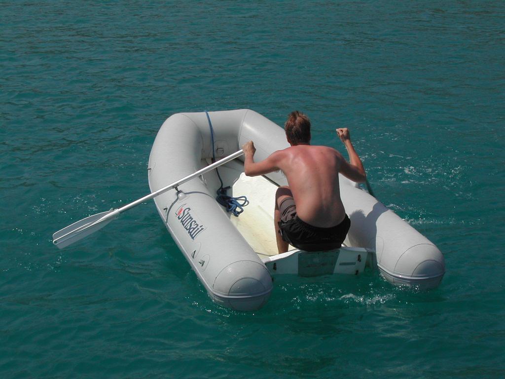 A man rows a small inflatable boat on clear turquoise water, using a single oar.
