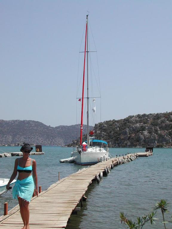 A woman in a swimsuit walks on a wooden dock, with a sailboat moored at the end.