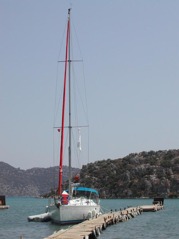 A sailboat with a red mast is docked at a wooden pier, surrounded by calm water and rocky hills.