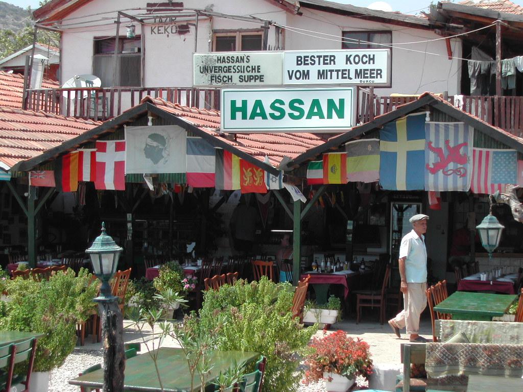 A restaurant named "Hassan" with international flags, outdoor seating, and a man in light clothing walking past.