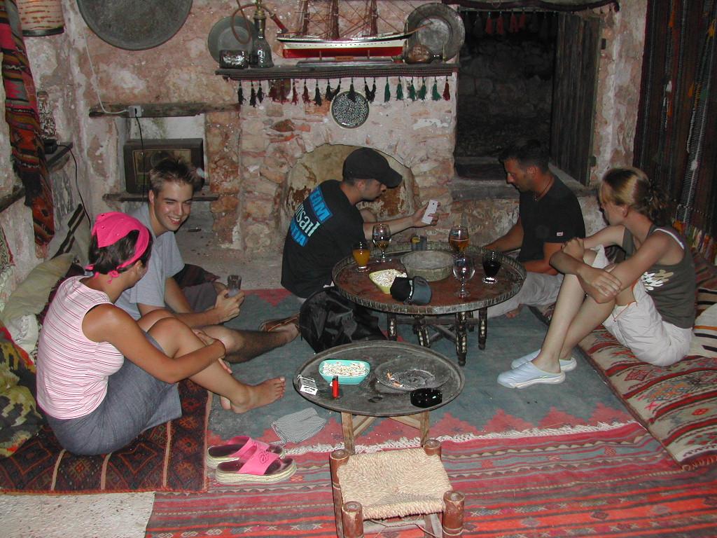 A group of five people sits on cushions around a low table, talking, laughing, and playing cards.