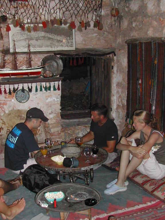 Three people sit on cushions around a low table, drinking and talking in a rustic, decorated room.