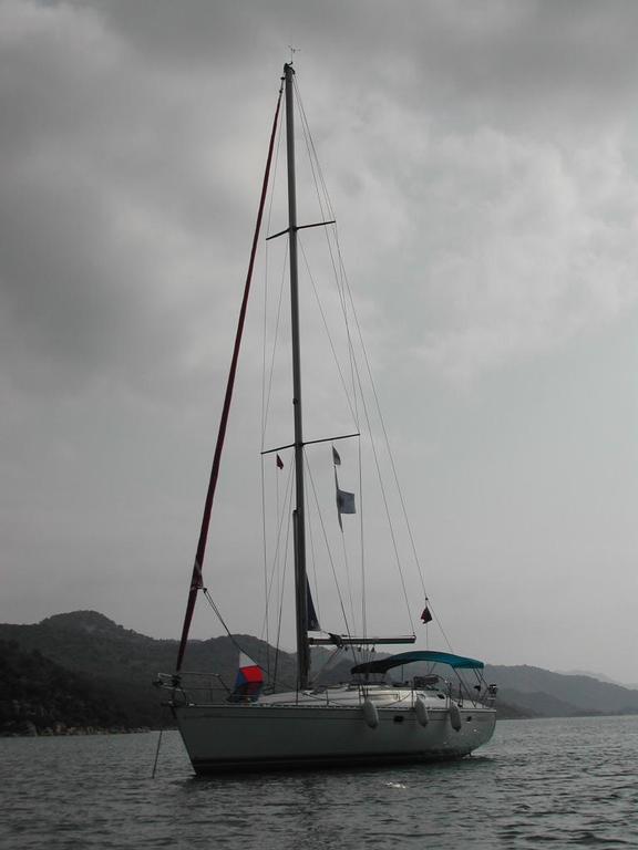 A sailboat with a tall mast floats on calm water, with hills and a cloudy sky in the background.