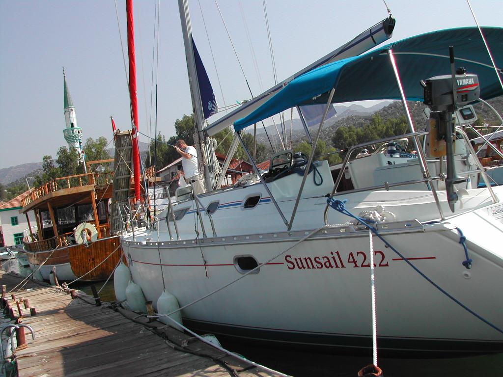 A man stands on a white sailboat docked at a wooden pier, adjusting something near the mast.