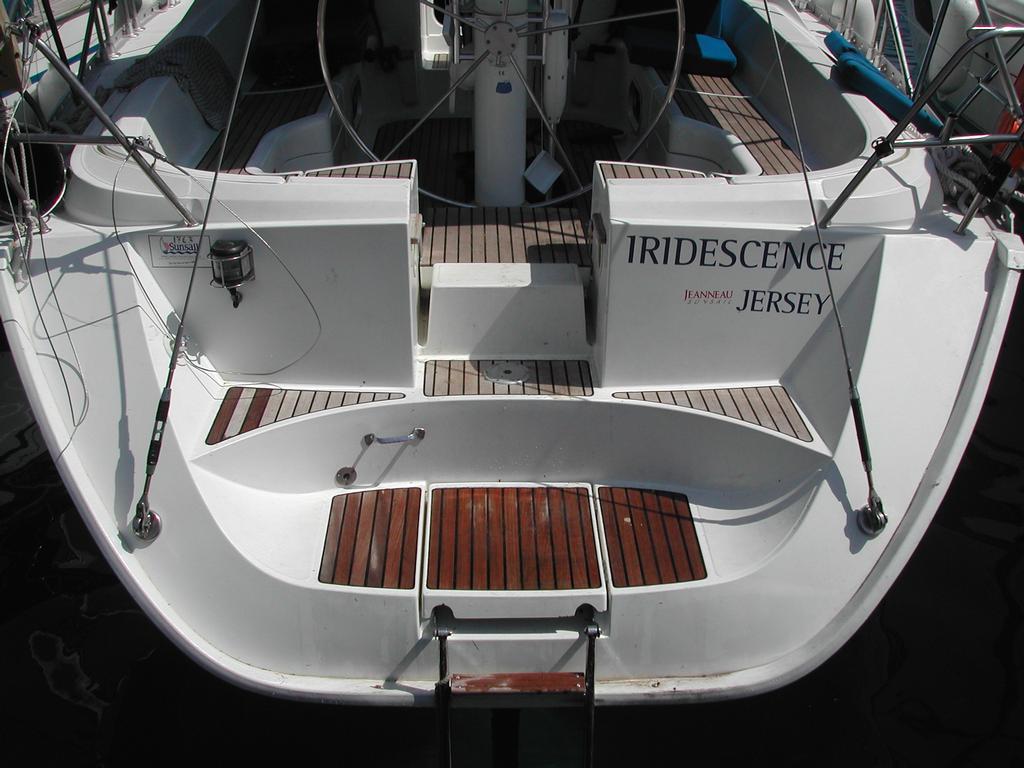 The back of a white sailboat named "Iridescence" with wooden steps, fishing rods, and seating in the cockpit.