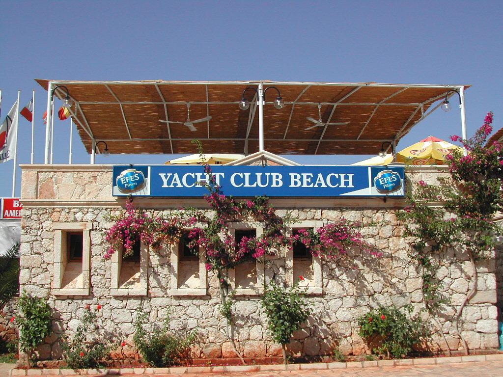 A stone building with a "Yacht Club Beach" sign, covered patio, and flowering plants in Kalkan, Turkey.
