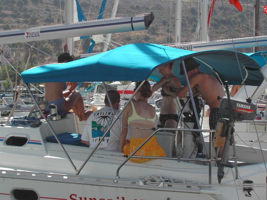 A group of people relax on a sailboat under a blue canopy in a marina.