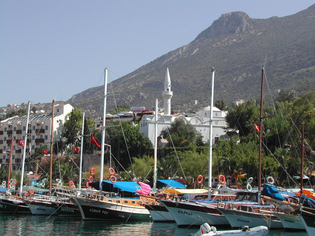 Several sailboats with Turkish flags docked in a harbor, with a white mosque and mountains in the background.