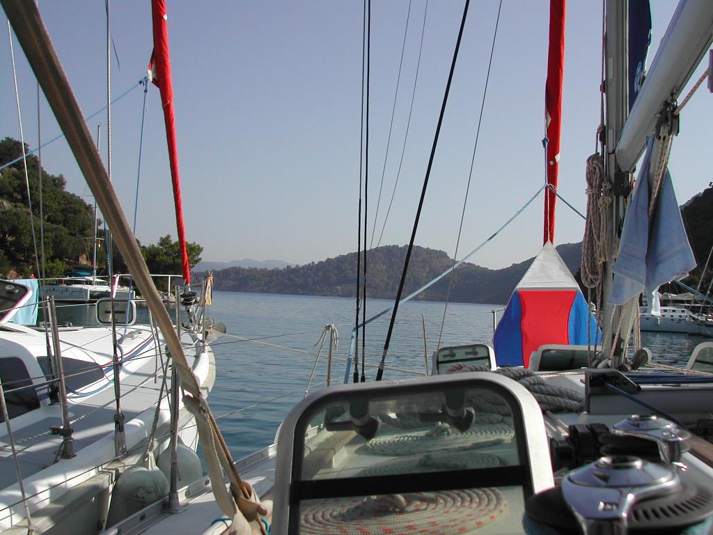 View from a sailboat docked in a calm bay, with other boats nearby and hills in the background.