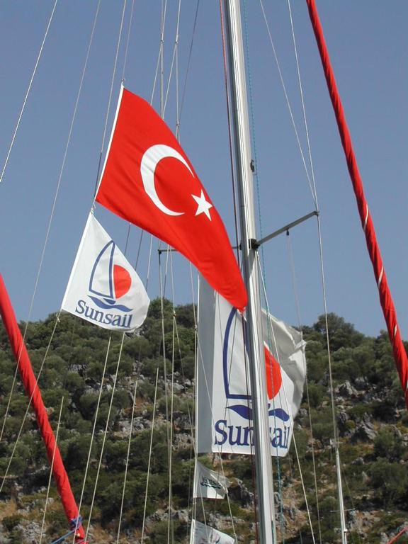 A sailboat mast with a Turkish flag and Sunsail banners, set against a hillside with green vegetation.