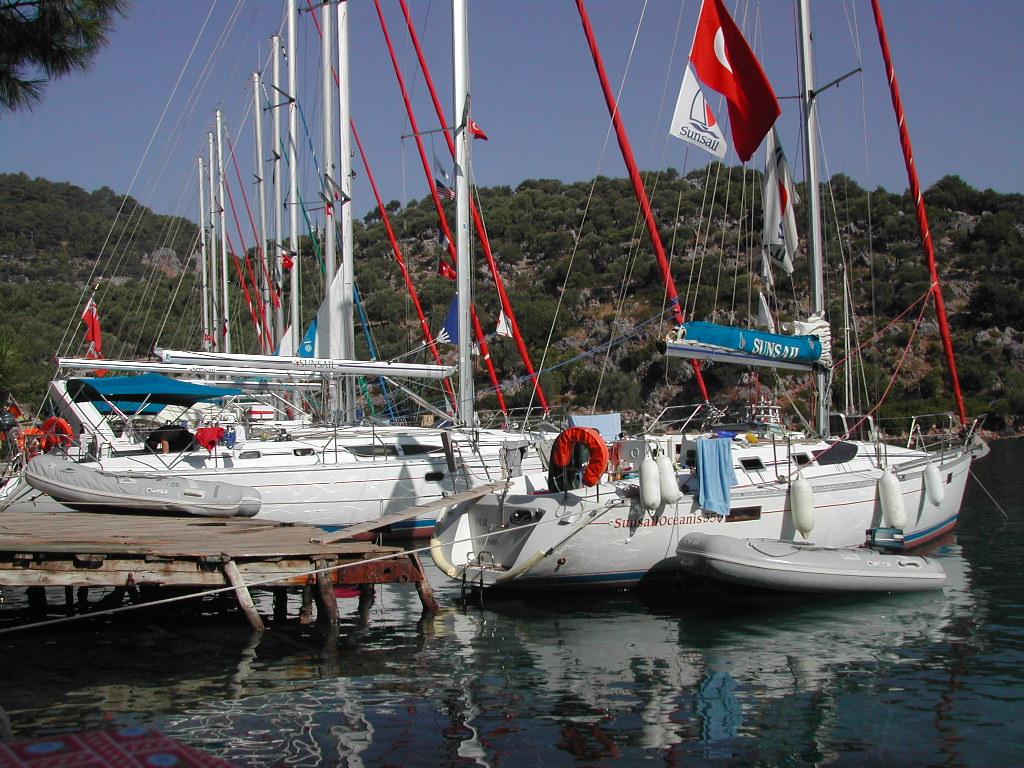 Several sailboats with red and white flags are docked at a wooden pier, with inflatable dinghies floating nearby.
