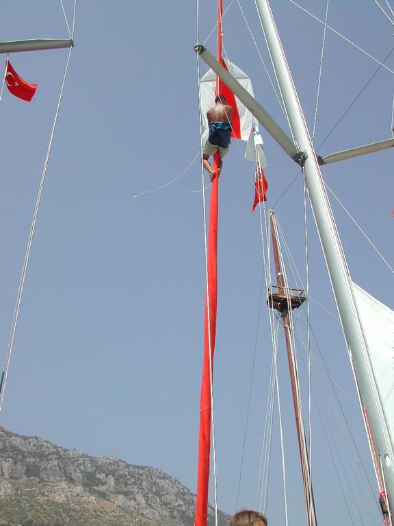 A person climbs a sailboat mast, holding onto a red flag, with mountains in the background.