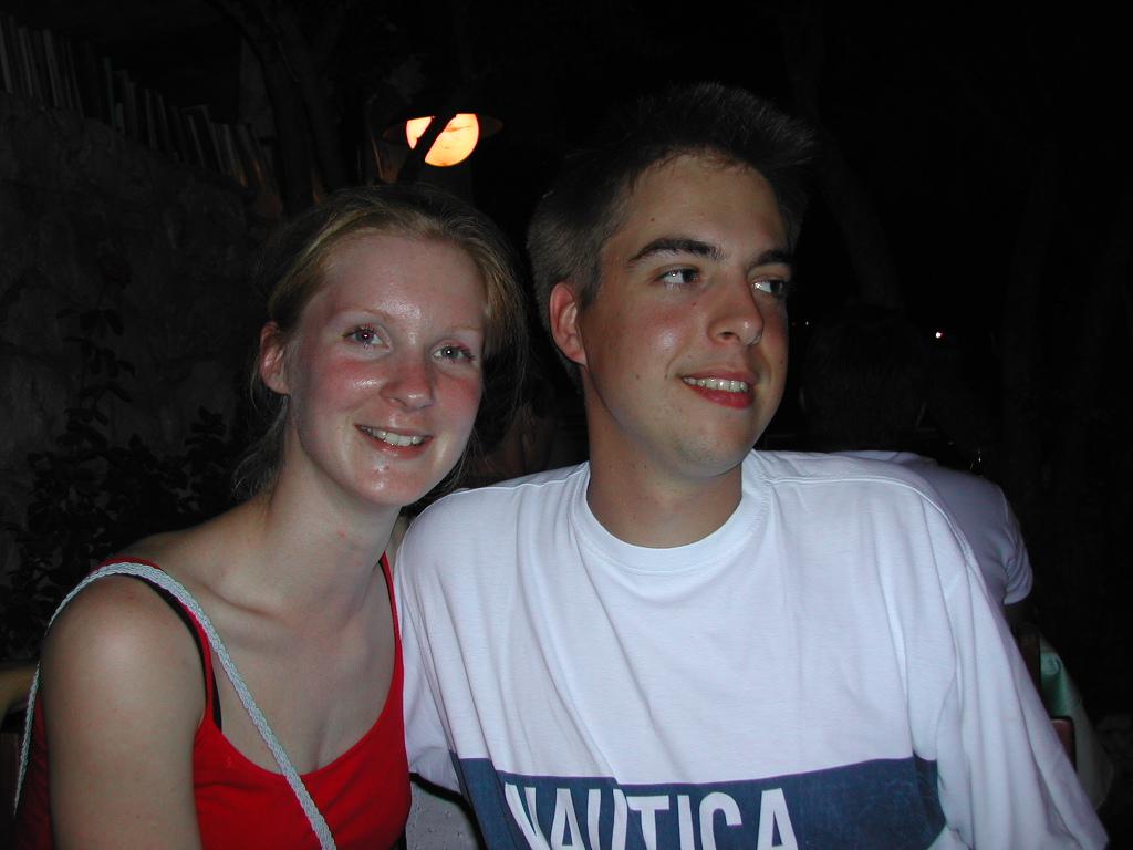 A smiling woman in a red top and a man in a white shirt sit together at night.