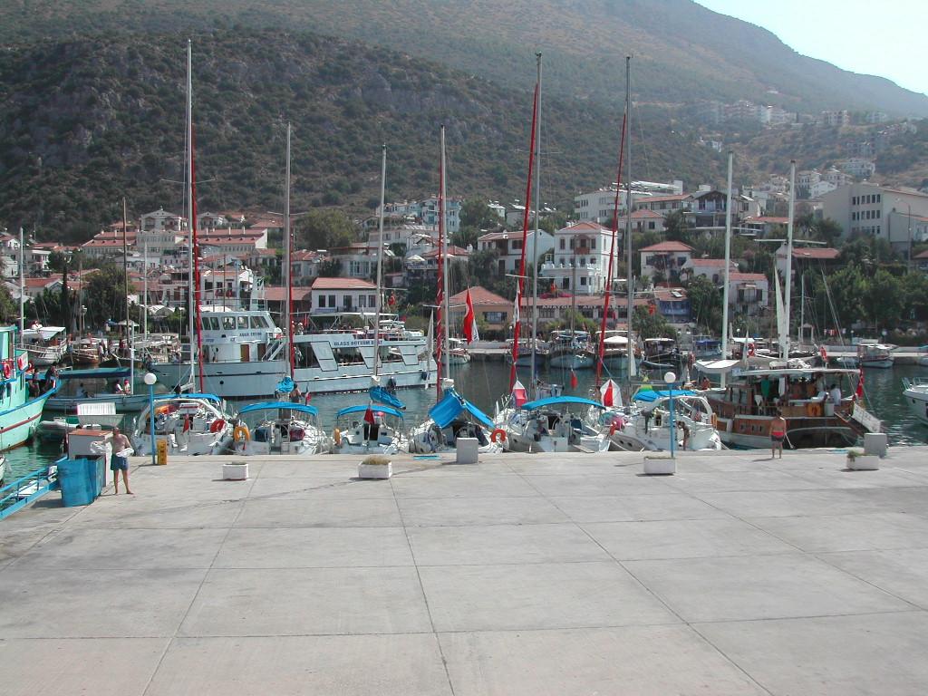 A marina in Kas, Turkey, with several sailboats docked and people walking along the pier.