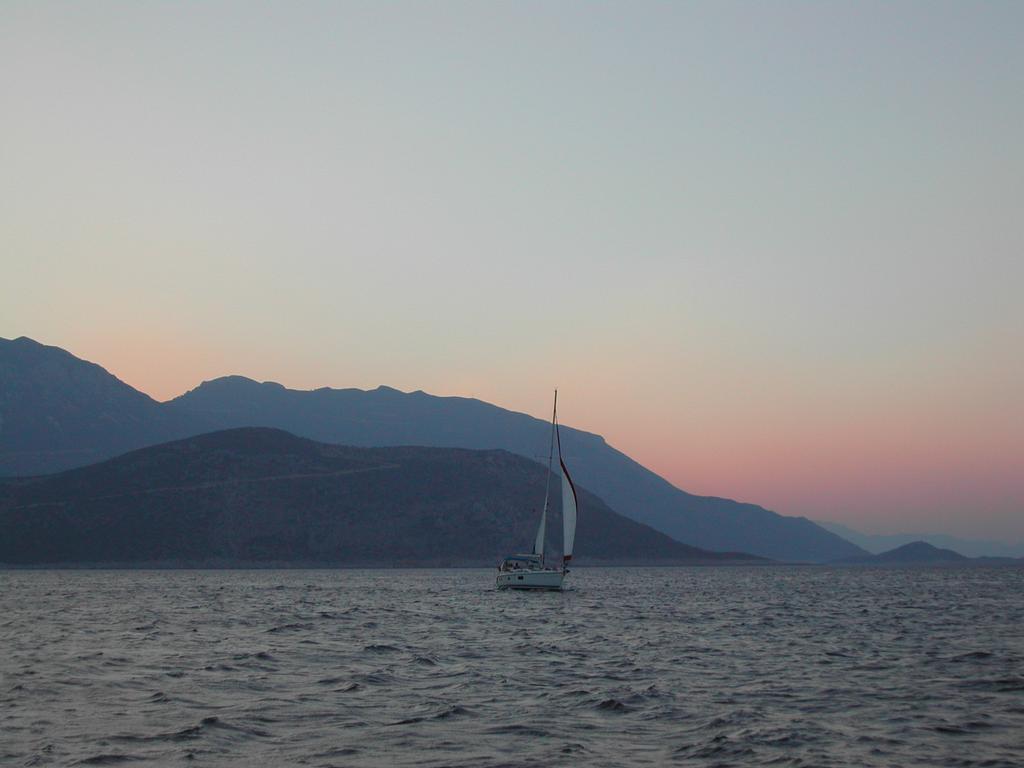 A sailboat moves across the water with distant mountains in the background during sunset.