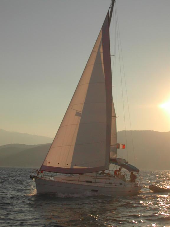 A sailboat moves through the water at sunset, with people on board and distant mountains in the background.