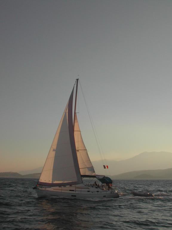 A sailboat with raised sails moves through the water at sunset, towing a small dinghy behind it.