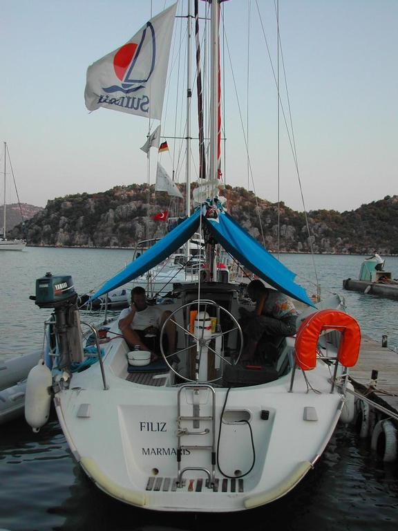A sailboat named "Filiz" docked at a marina, with two people sitting under a blue tarp on board.