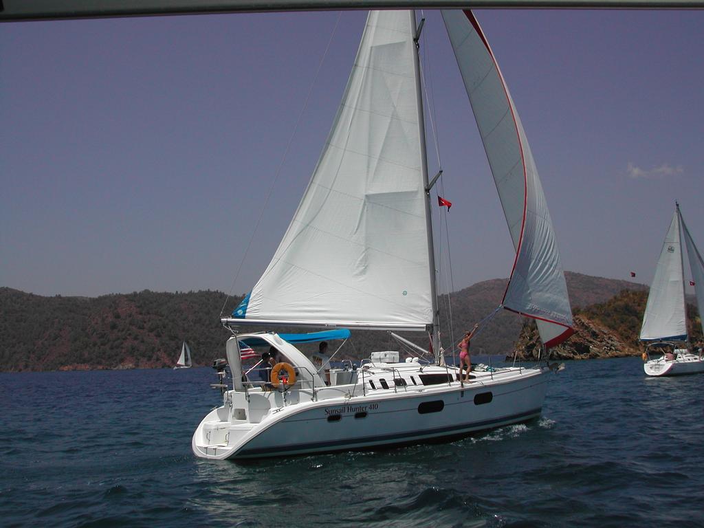 A sailboat with white sails moves through the water, with a person standing near the front adjusting a rope.