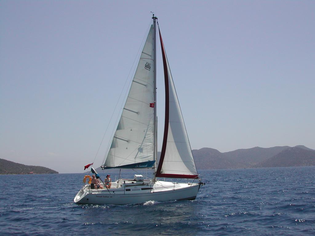 A sailboat with a tall white sail moves through the water, with people on board.