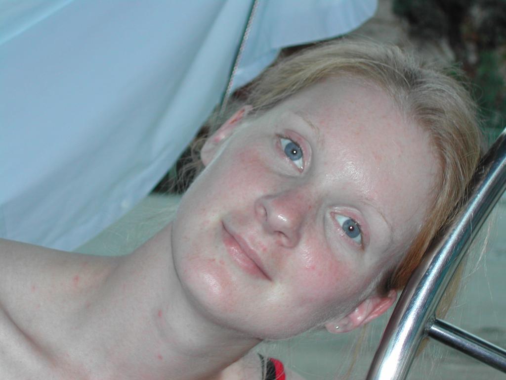 A woman with light skin and blonde hair leans her head on a metal railing, looking directly at the camera.