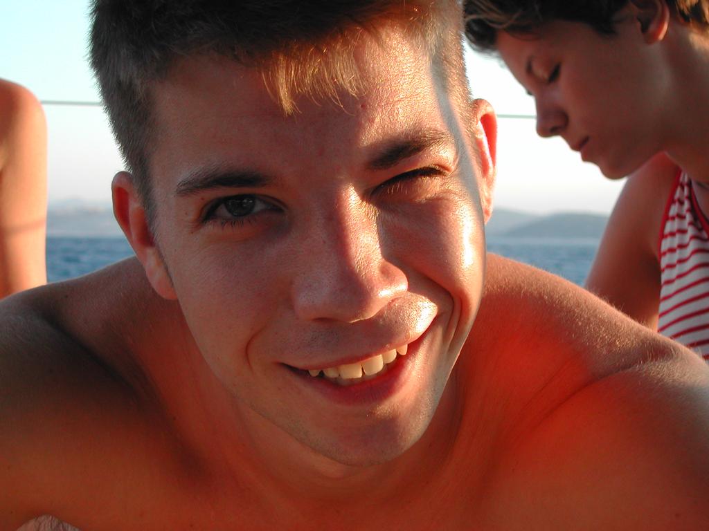 A man smiles and winks at the camera, while two people sit in the background near the water.