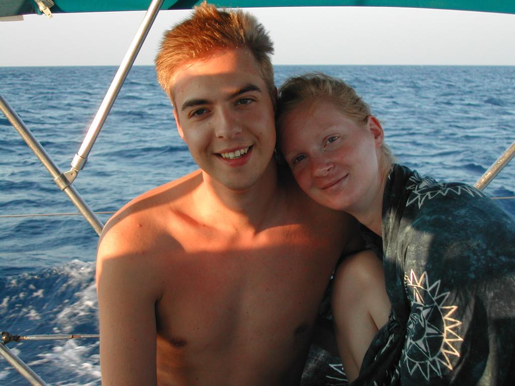 A smiling couple sits close together on a boat with the ocean in the background.