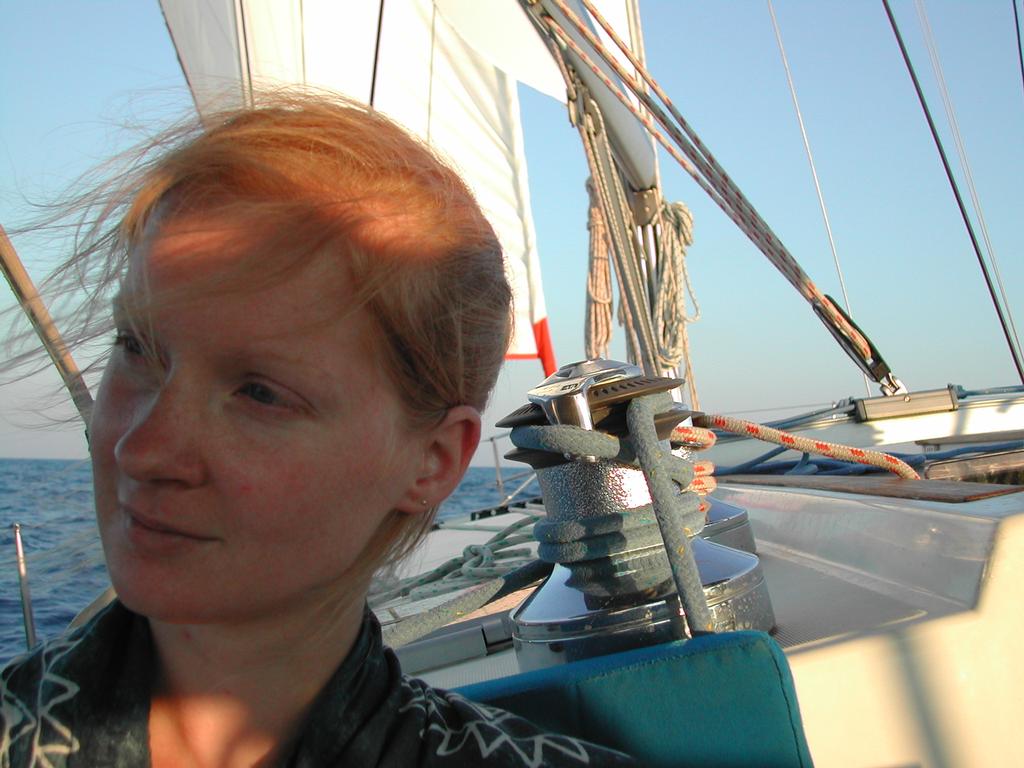 A woman with windblown hair sits on a sailboat, with ropes and sails visible in the background.