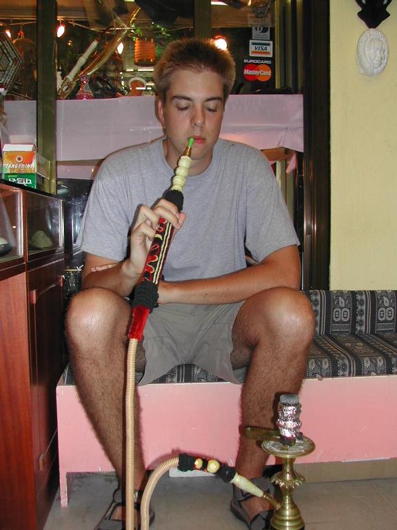 A man in a gray shirt sits on a cushioned bench, smoking a hookah in an indoor setting.