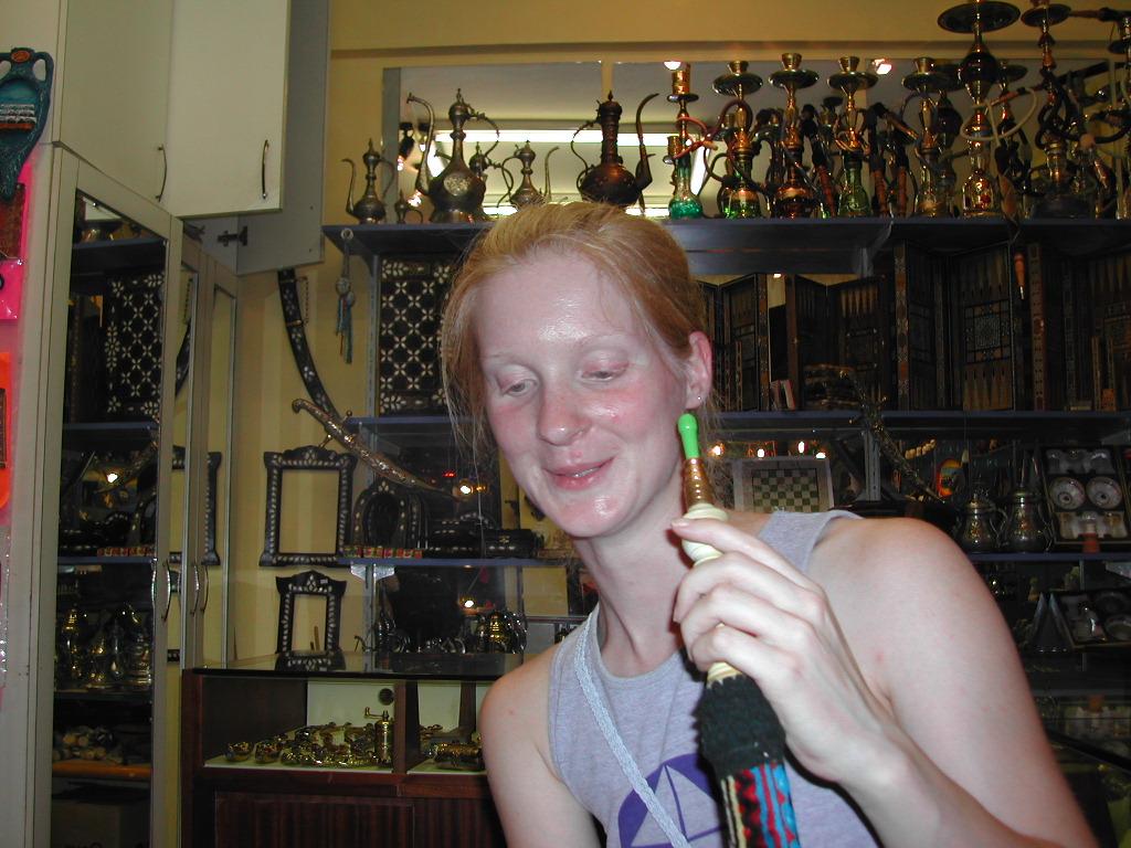 A woman holds a hookah pipe and smiles inside a shop filled with decorative items and hookahs.