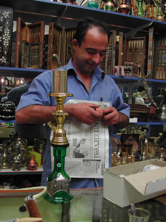 A man in a blue shirt smiles while preparing a hookah in a shop filled with decorative items.