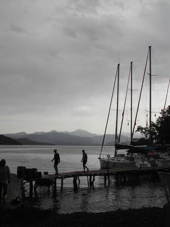 Two people walk on a wooden dock toward sailboats, with mountains and water in the background.