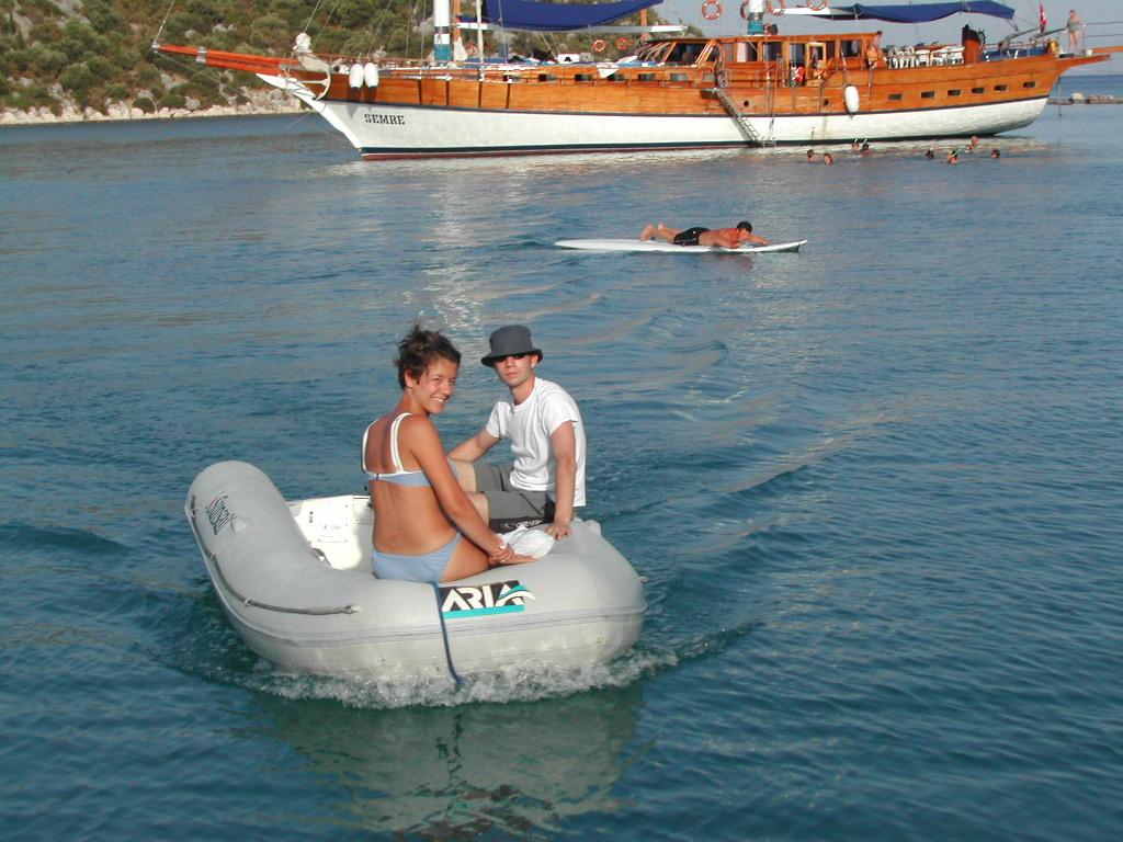 Two people ride a small inflatable boat on the water, with a large wooden ship and swimmers in the background.