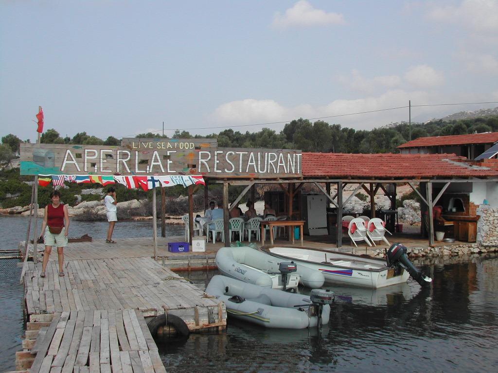 A floating seafood restaurant with a wooden dock, boats, and people walking and sitting at tables.