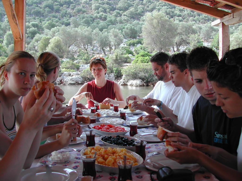 A group of people sits at an outdoor table, eating bread and other food while drinking tea.