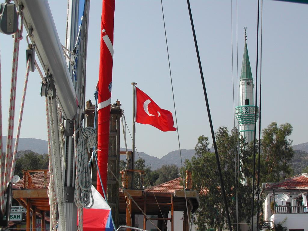 A Turkish flag waves near a sailboat, with a mosque minaret and buildings in the background.