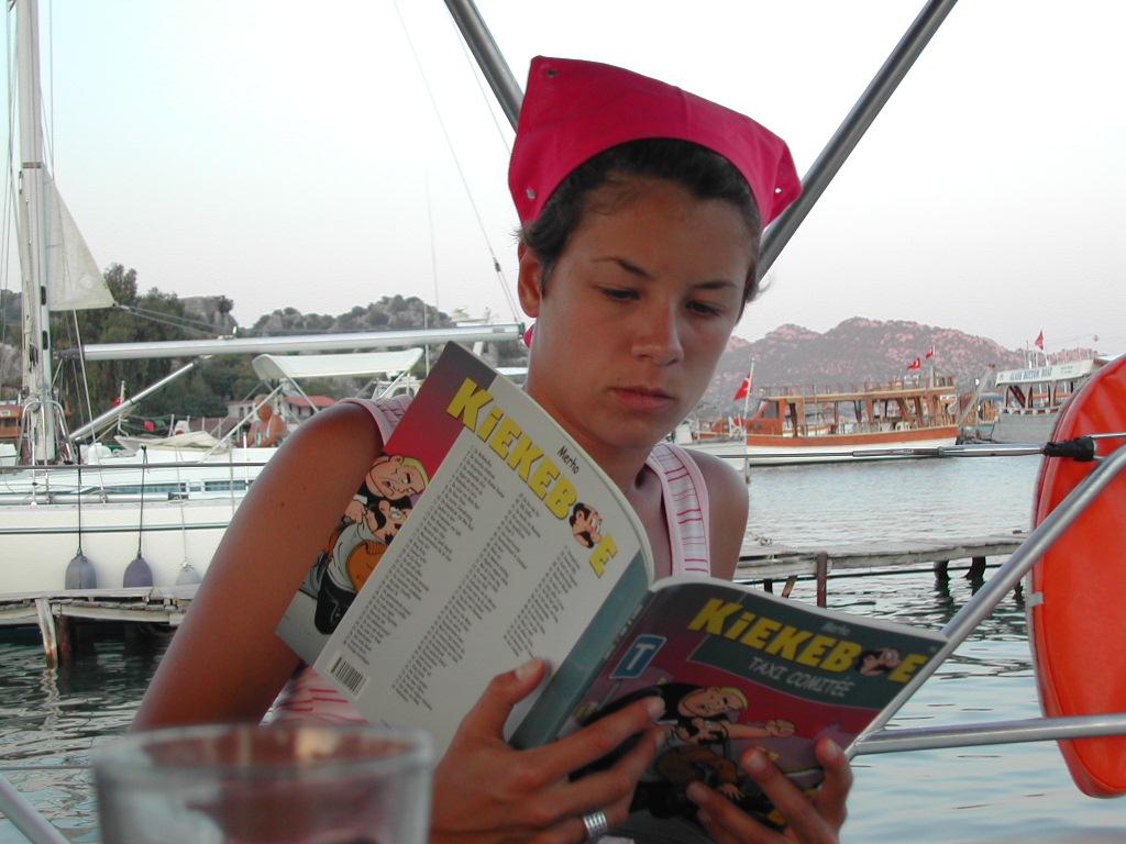 A young person wearing a red headscarf reads a comic book while sitting on a boat.